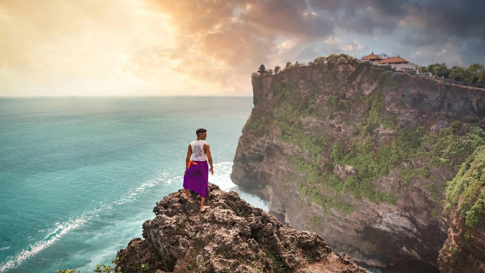Traveller in a sarong on a clifftop rock facing Uluwatu Temple cliffs at golden sunset