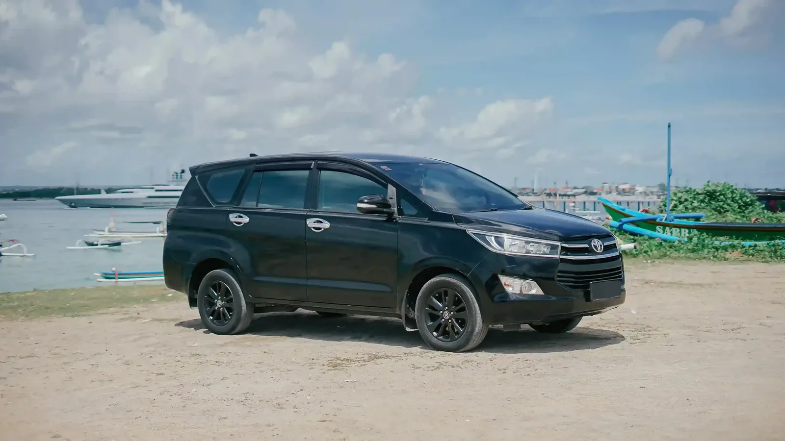 Black Toyota Innova tour car parked on a sandy shore lot with fishing boats in Bali