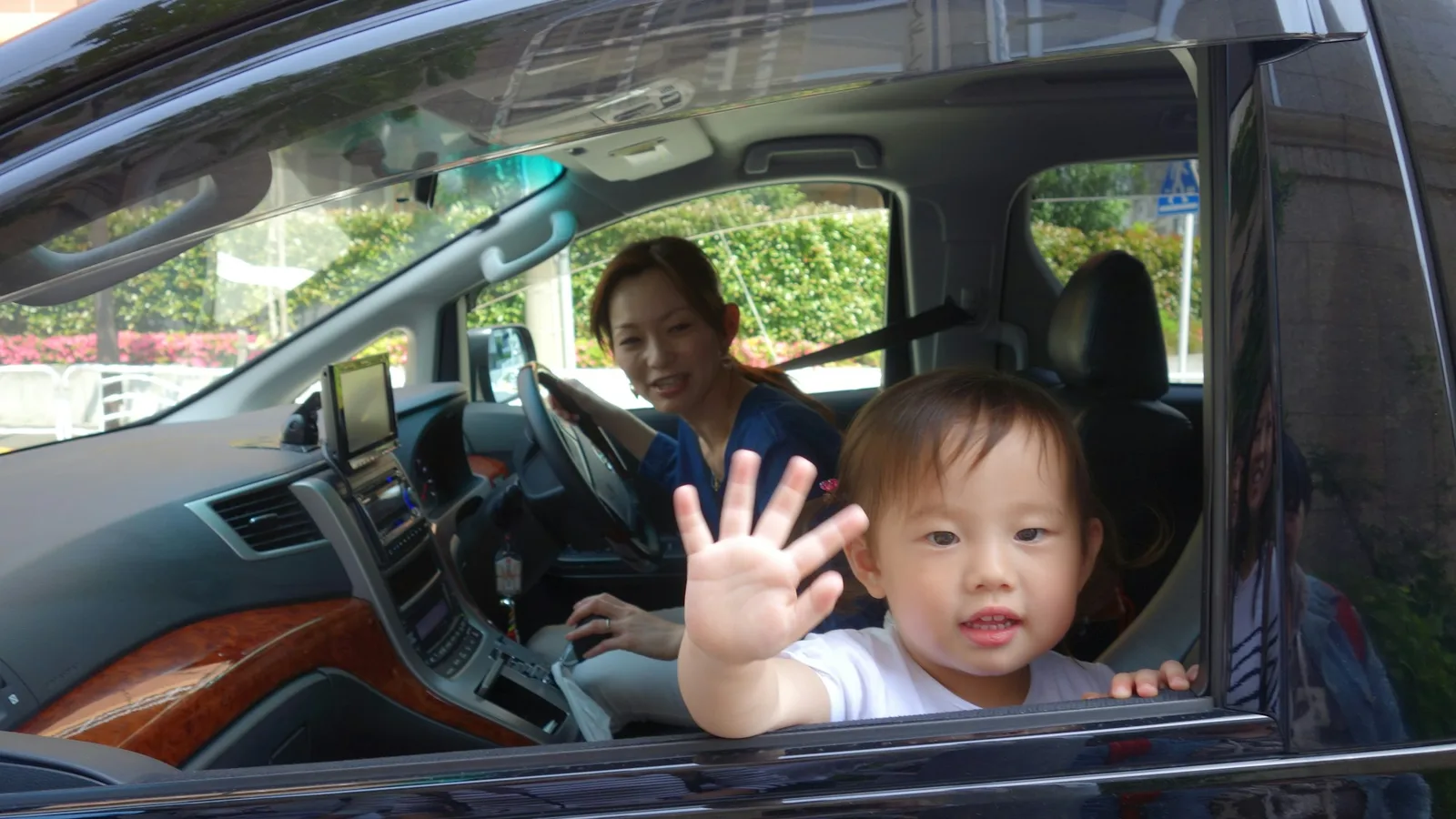 Toddler waving from the window of a family MPV while a smiling mother sits at the wheel